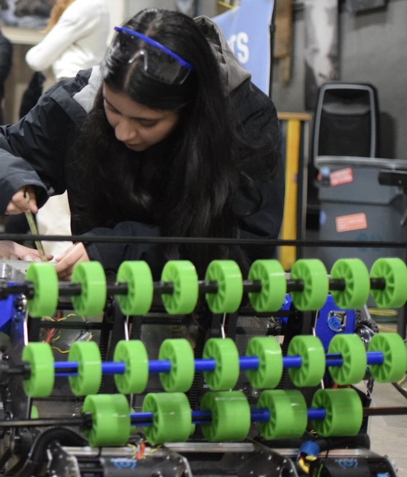 Afrah Nazar leaning over a robot mechanism with a tool, safety glasses pushed up on her head